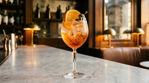 Grand verre à vin rempli de glace et d'un spritz orange vif pétillant, demi-rondelle d'orange et olive verte sur le bord, sur un comptoir de marbre veiné dans un bar chic