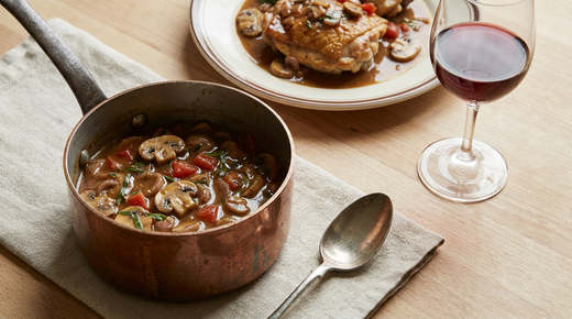 Petite casserole en cuivre de sauce chasseur brune aux champignons, échalote, tomate et estragon, cuisse de poulet sautée nappée, verre de vin rouge, plan de travail en bois