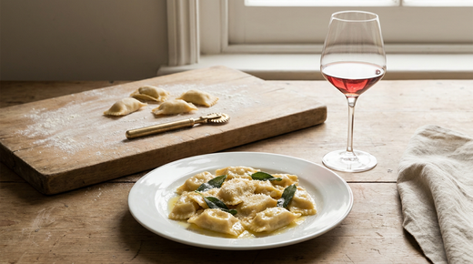 Assiette d'agnolotti del plin au beurre et à la sauge, parmesan, agnolotti crus sur une planche farinée avec une roulette, verre de vin rouge, plan de travail en bois