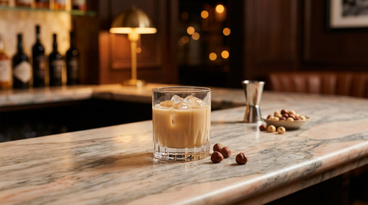 Nutty Irishman crémeux caramel sur glace dans un verre bas, noisettes entières posées à côté, comptoir de marbre Fior di Pesco