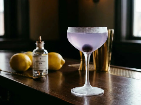 Aviation violet pâle en coupette avec cerise au fond, citrons et bouteille de gin en arrière-plan sur un bar sombre