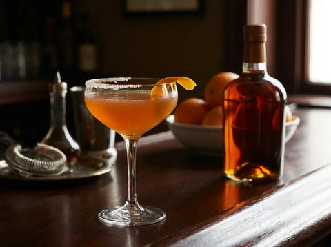 Sidecar ambré en coupette à demi-croûte de sucre avec zeste d'orange, bouteille d'eau-de-vie et oranges sur bar sombre