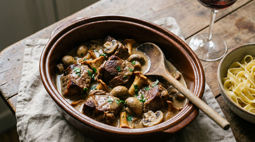 Plat en terre vernissée de sauté de porc aux champignons, morceaux d'épaule dorés dans une sauce crémeuse brune, champignons de Paris et girolles, persil, cuillère en bois à plat, bol de tagliatelles, verre de vin rouge, table en bois