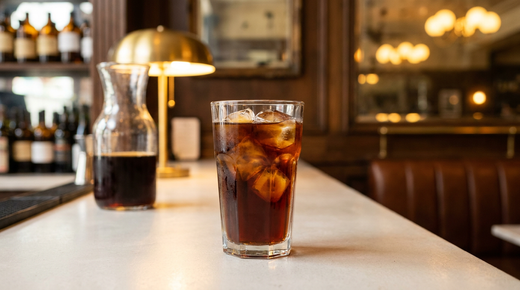 Cold brew brun profond et limpide dans un grand verre à glaçons nets, carafe floue derrière, comptoir de béton clair