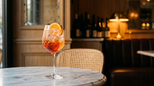 Grand verre à vin rempli de glace et d'un apéritif orange-rouge translucide, demi-rondelle d'orange sur le bord, sur une table de bistrot en marbre au crépuscule