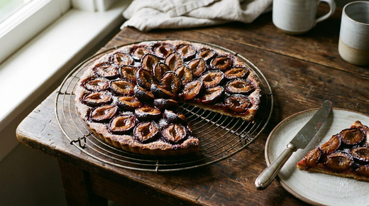 Tarte aux prunes dorée sur une grille, demi-prunes violettes et dorées rangées serrées face coupée vers le haut, jus caramélisé au fond des creux, pâte brisée brune sur le pourtour, sucre glace, table de bois patiné