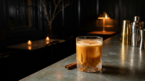 Verre bas d'un cocktail au potiron orangé et crémeux sur un gros glaçon, bâton de cannelle posé, sur le comptoir de zinc d'un bar dans la pénombre, bougies allumées au fond
