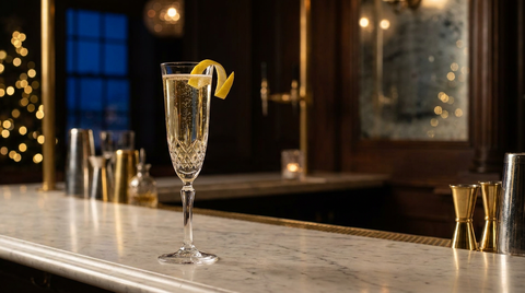Flûte en cristal d'un cocktail pétillant doré lumineux, long zeste de citron arqué sur le bord, sur un comptoir de marbre, bokeh doré et bleu de minuit à la fenêtre