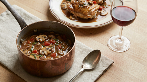 Petite casserole en cuivre de sauce chasseur brune aux champignons, échalote, tomate et estragon, cuisse de poulet sautée nappée, verre de vin rouge, plan de travail en bois