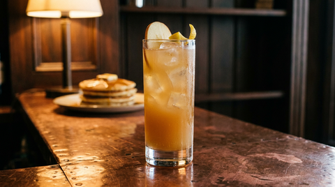 Cocktail à la pomme doré trouble dans un grand verre sur glaçons, fine lamelle de pomme et zeste de citron, pile de crêpes floue derrière, comptoir de cuivre, lumière chaude