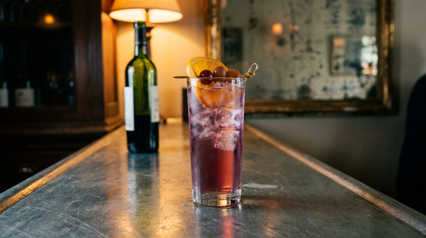 Cocktail au vin nouveau rouge vif dans un grand verre plein de glaçons, tranche d'orange et grains de raisin, comptoir de zinc brossé, lumière chaude de bistrot de novembre