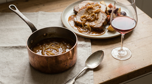 Petite casserole en cuivre de sauce lyonnaise brune aux oignons fondus, tranches de rôti de porc nappées, verre de vin rouge, plan de travail en bois