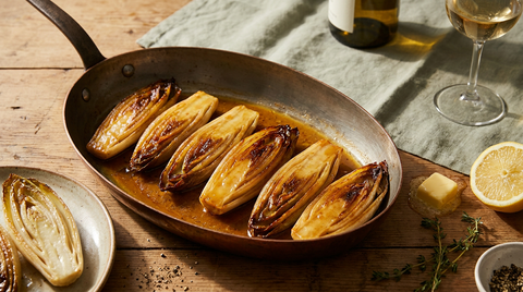 Endives braisées dorées et laquées dans une poêle en cuivre, glaçage ambré, citron et thym à côté