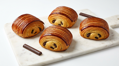Quatre pains au chocolat entiers sur une planche de marbre blanc, dessus laqué brun caramel aux stries fines et régulières, feuilletage enroulé visible en bout avec le bâton de chocolat, deux bâtons de chocolat posés à côté