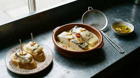 Bacalao al pil-pil dans une cazuela en terre, morceaux de morue blanche nappés d'une sauce ivoire brillante, lamelles d'ail confit et rondelle de piment, deux morceaux servis sur baguette avec des piques, passoire métallique posée à côté