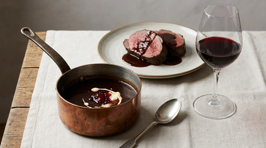 Petite casserole en cuivre de sauce chevreuil acajou avec gelée de groseille et crème, tranches de chevreuil rôti nappées, verre de vin rouge, plan de travail en bois