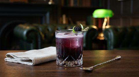 Cocktail d'Halloween violet foncé aux mûres et au gin dans un verre bas taillé, gros glaçon, col de mousse pâle, mûre et zeste de citron vert, sur une table de noyer