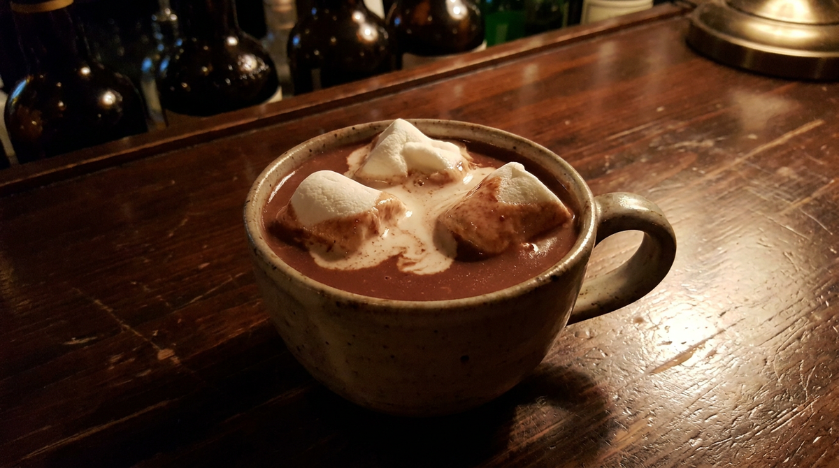 Tasse épaisse remplie d'un chocolat chaud brun foncé surmonté de deux guimauves blanches à demi fondues, comptoir de bois ciré sombre, lumière chaude et basse