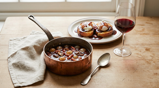 Sauteuse en cuivre de sauce bourguignonne au vin rouge avec petits oignons glacés, champignons et lardons, œufs pochés en meurette sur pain grillé, verre de vin rouge, plan de travail en bois