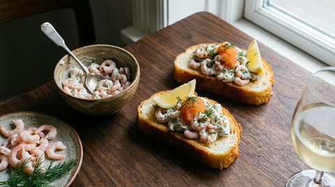 Deux toasts Skagen sur une planche en bois sombre, pain doré au beurre couvert de petites crevettes roses entières liées d'une mayonnaise à l'aneth, œufs de lump orange, aneth frais et quartier de citron