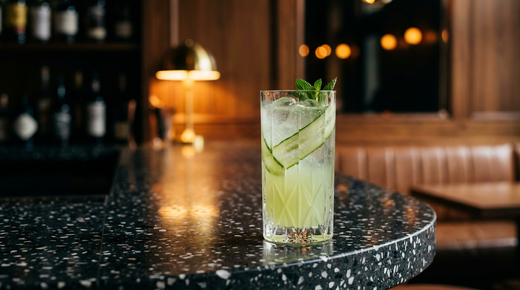 Cucumber Cooler vert très pâle et pétillant dans un verre haut, rubans de concombre enroulés et brin de menthe, comptoir de terrazzo anthracite