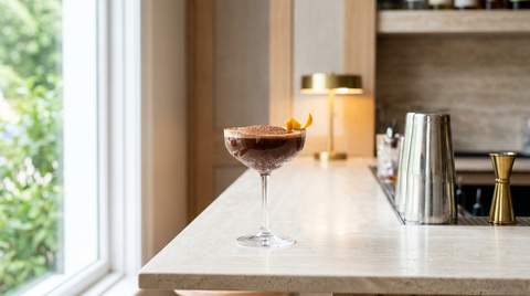 Coupe en cristal d'un cocktail chocolat-orange brillant saupoudré de cacao, zeste d'orange sur le bord, sur un comptoir de travertin clair, lumière de matin de printemps