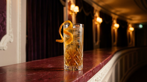 Rivesaltes tonic en verre haut de cristal, glace claire et long zeste d'orange, sur la tablette de marbre du foyer d'un théâtre aux appliques dorées