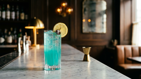 Grand verre de glace et de cocktail bleu turquoise lumineux, rondelle de citron jaune sur le bord, sur un comptoir de marbre veiné dans un bar chic à la lumière du soir