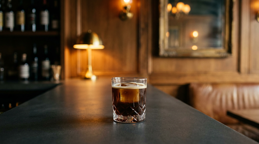 Carajillo brun café sur un gros glaçon dans un petit verre, fine ligne de crème en surface, comptoir de béton anthracite