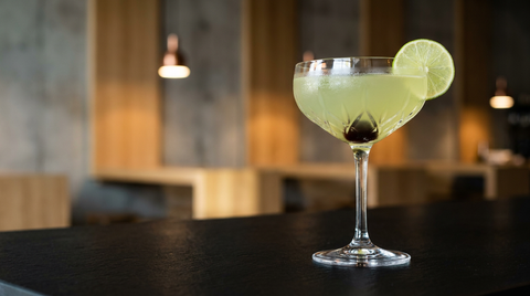 Closing Argument en coupe de cristal sur un comptoir d'ardoise noire dans un bar à vins moderne, robe vert pâle, cerise au fond et rondelle de citron vert