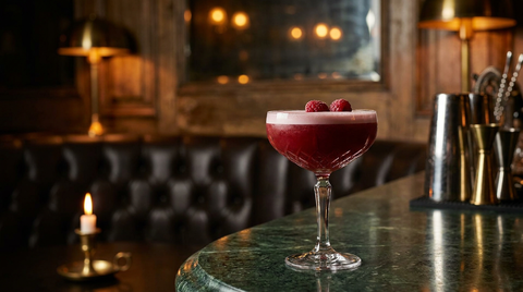 Coupe en cristal d'un cocktail aux fruits rouges sous une mousse pâle, framboises posées dessus, sur un marbre vert, bougie allumée et banquette de cuir au fond