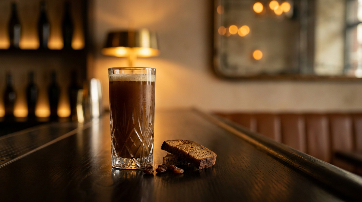 Kvass brun trouble avec un col de mousse fauve dans un verre haut, pain de seigle grillé et raisins secs à côté, comptoir de chêne des marais