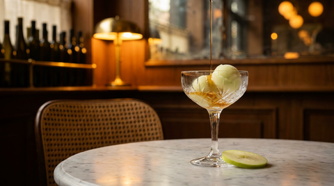 Coupe en cristal de sorbet pomme verte arrosé d'un filet d'armagnac ambré, lamelle de pomme à côté, sur une table de bistrot en marbre au crépuscule