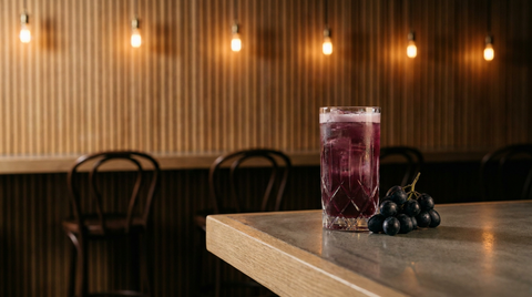Cocktail sans alcool au raisin frais dans un verre haut de cristal, robe pourpre à col rose, grappe de raisin noir, sur le comptoir de béton ciré d'un bar à vins moderne