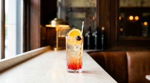 Grand verre de glace avec une boisson en dégradé du doré au rouge, rondelle d'orange et cerise sur le bord, paille en papier, dans la lumière claire du matin sur un comptoir clair