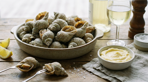 Bulots cuits en tas dans un plat de grès, quelques-uns décortiqués sur des fourchettes, mayonnaise maison, citron et verre de vin blanc