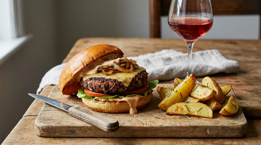 Burger végétarien maison, pain brioché, steak de haricots rouges et champignons grillé, cheddar fondu, oignons caramélisés, salade, tomate et sauce, pommes de terre rôties au romarin, couteau à plat, verre de vin rouge, planche en bois