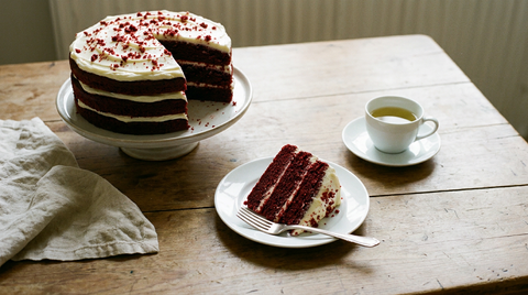 Red velvet cake à trois étages sur un présentoir, une part coupée montrant la mie rouge foncé fine et le glaçage blanc au fromage frais, miettes rouges sur le dessus, tasse de thé vert