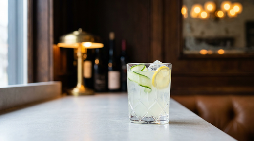 Limonade au concombre vert très pâle dans un verre droit sur glace, rubans de concombre et rondelle de citron, plan en pierre claire