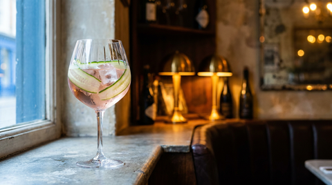 Cocktail rosé concombre dans un grand verre à pied, rubans de concombre, appui de fenêtre en pierre