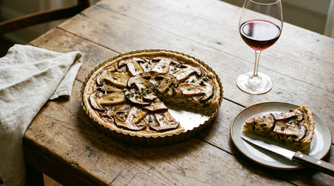 Tarte salée aux cèpes dans un moule cannelé, lamelles de cèpes dorées sur un appareil à la crème, une part coupée sur une assiette, couteau à plat, verre de vin rouge, table en bois