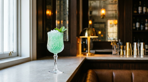 Verre à pied givré rempli de glace pilée et de liqueur de menthe vert clair, feuille de menthe posée dessus, sur un appui de fenêtre en pierre au bord d'un comptoir de bar