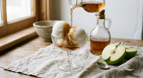 Coupe en verre à pied avec deux boules de sorbet blanc à la pomme arrosées d'un filet d'eau-de-vie ambrée, carafe d'alcool ambré et pomme verte coupée sur une nappe de lin