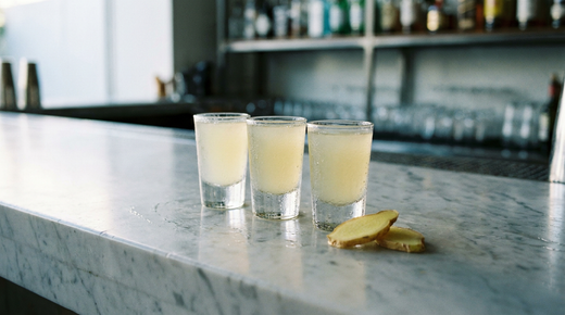 Trois petits verres à shooter alignés contenant un liquide jaune pâle trouble très froid, rondelles de gingembre à côté, comptoir de marbre clair, lumière vive et nette