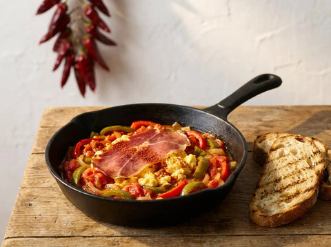 Piperade basque dans sa poêle : poivrons rouges et verts confits aux tomates, œufs crémeux à peine pris et tranche de jambon de Bayonne