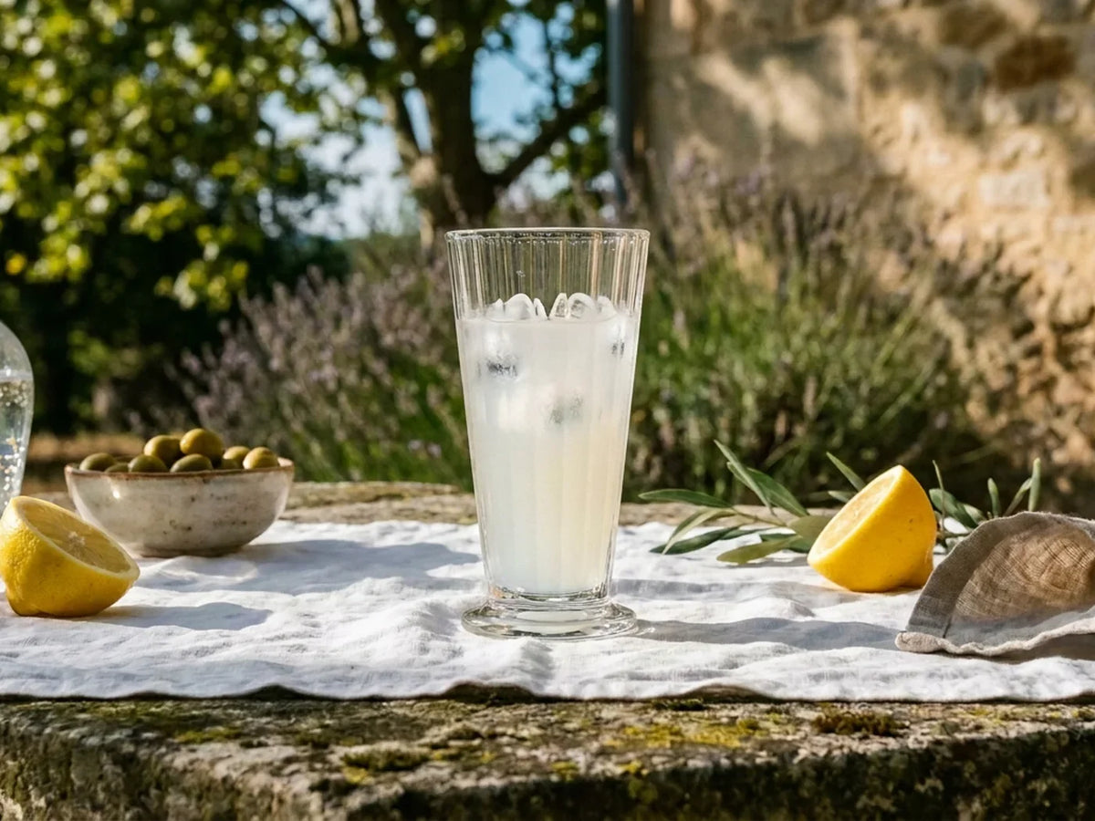 Verre à pastis côtelé sur une table de pierre nappée de lin, coupelle d'olives et citrons au soleil