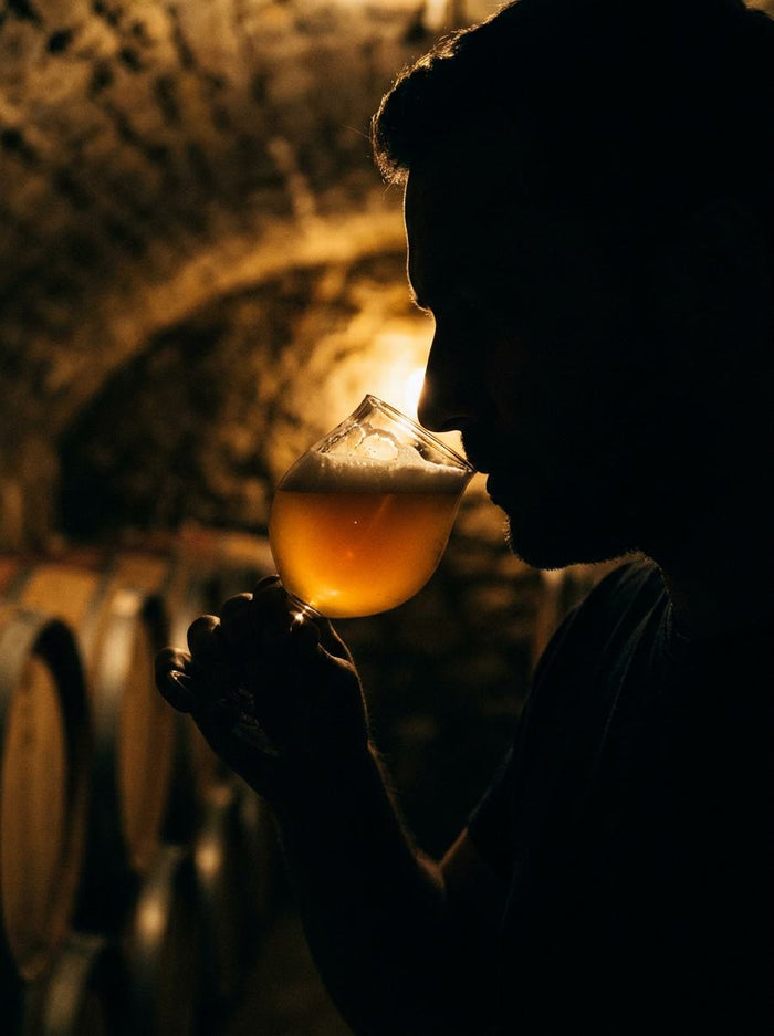 Silhouette d'un dégustateur au verre de bière dorée devant les fûts de la cave