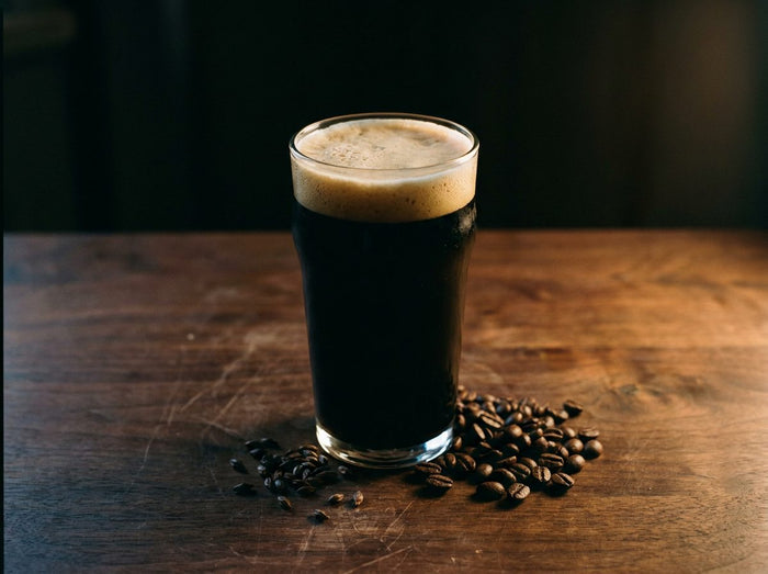 Verre de bière noire opaque à mousse beige, grains de café torréfiés sur une table de chêne
