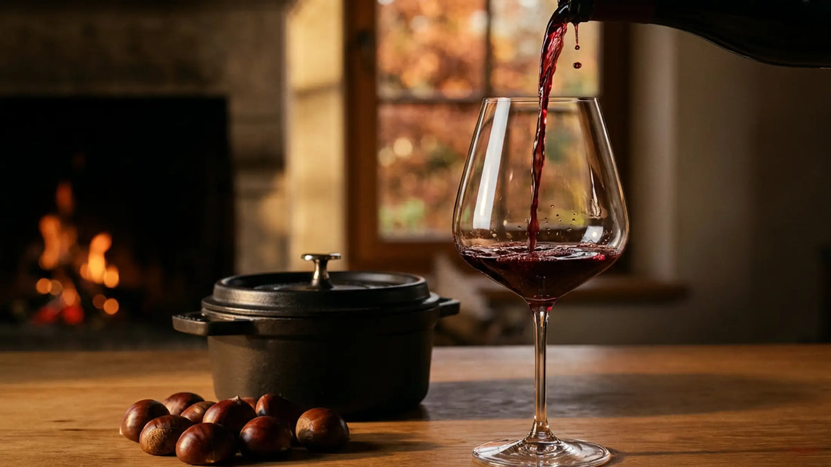 Vin rouge versé dans un grand verre devant une cheminée allumée, cocotte en fonte et châtaignes sur une table de chêne, feuillage d'automne à la fenêtre