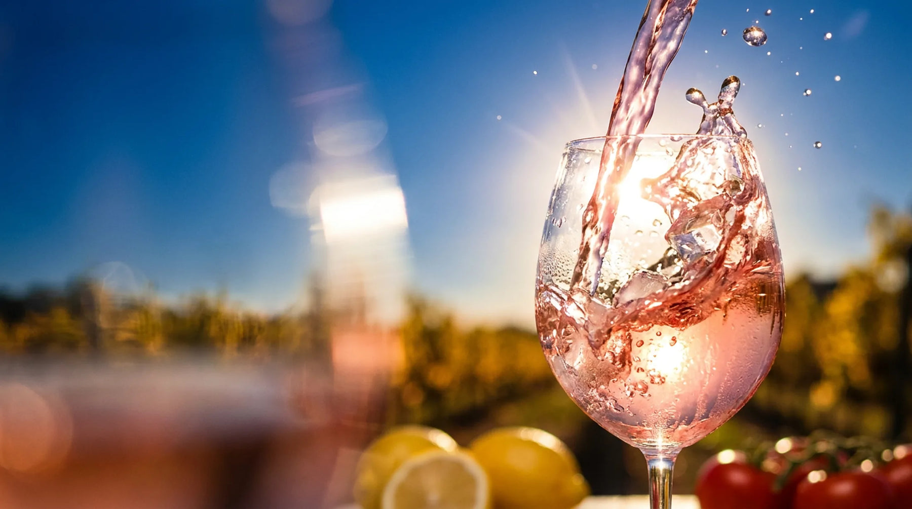 Vin rosé versé dans un grand verre à contre-jour, éclaboussures et glaçons figés, ciel bleu et vignes en fond